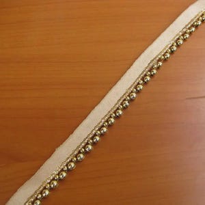 May include: Beige ribbon with gold-colored beaded trim. The ribbon is angled across a wooden surface. The trim features small, round beads and a thinner gold thread. This decorative trim is suitable for various craft projects.