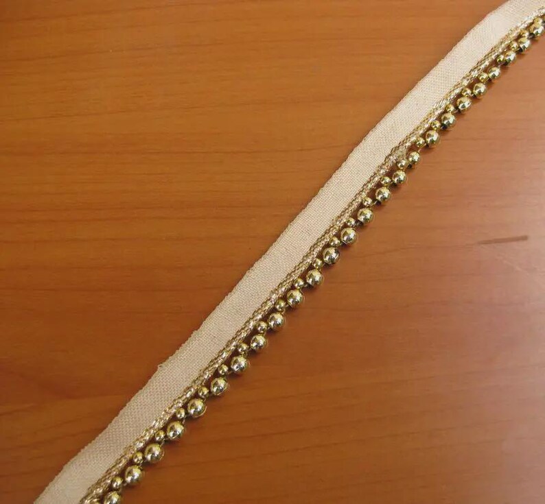 May include: Beige ribbon with gold-colored beaded trim. The ribbon is angled across a wooden surface. The trim features small, round beads and a thinner gold thread. This decorative trim is suitable for various craft projects.