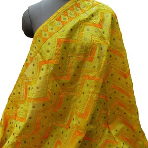 May include: A yellow textile with orange geometric patterns and small, reflective embellishments. The fabric is draped over a mannequin, showcasing the intricate embroidery and design. The textile has a vibrant, eye-catching appearance.