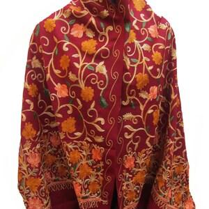 Puede incluir: Un chal de color granate con intrincados bordados florales en tonos naranja y amarillo. El chal tiene un borde con flecos.