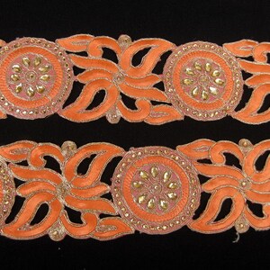 Op de afbeelding: Oranje en gouden trim met een bloemenpatroon en parelmoeraccenten. De trim is gemaakt van stof en heeft een delicaat, ingewikkeld patroon.