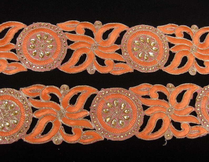 Op de afbeelding: Oranje en gouden trim met een bloemenpatroon en parelmoeraccenten. De trim is gemaakt van stof en heeft een delicaat, ingewikkeld patroon.