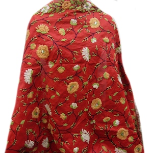 Op de afbeelding: Een rode sjaal met een geborduurd bloemmotief. De sjaal heeft gele, witte en groene bloemen met donkergroene ranken. De stof is gedrapeerd en toont de ingewikkelde borduursels en levendige kleuren. Dit textiel is geschikt voor diverse gelegenheden.