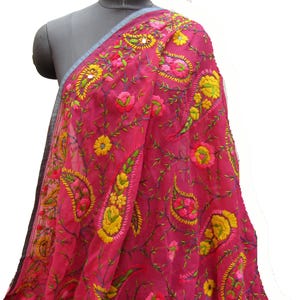 May include: A bright pink embroidered scarf featuring yellow, green, and pink floral and paisley designs. The scarf is displayed on a mannequin, highlighting its delicate, sheer fabric and detailed needlework.