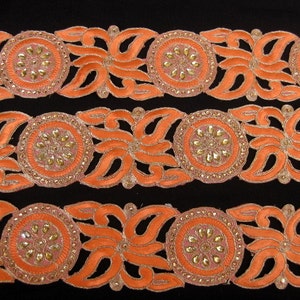 Op de afbeelding: Oranje en gouden trim met een bloemenmotief en parelmoeraccenten. De trim is gemaakt van stof en heeft een gegolfde rand.
