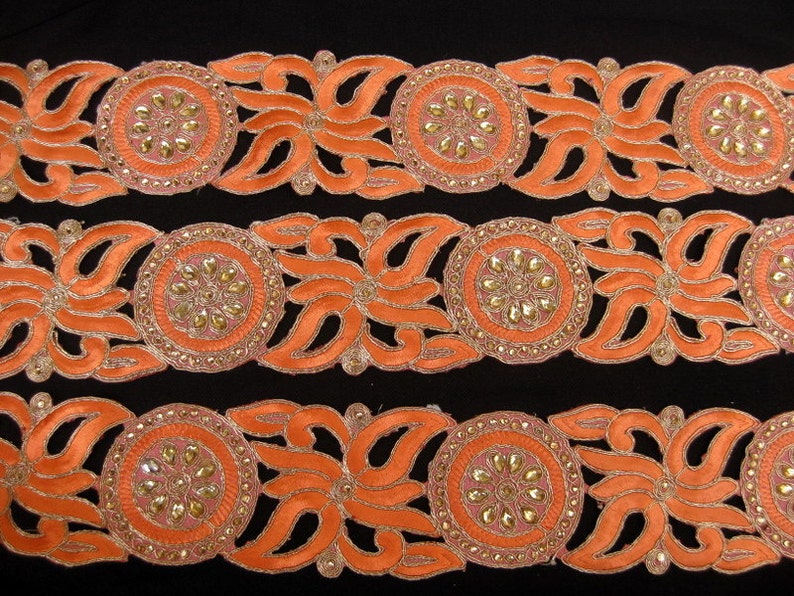 Op de afbeelding: Oranje en gouden trim met een bloemenmotief en parelmoeraccenten. De trim is gemaakt van stof en heeft een gegolfde rand.