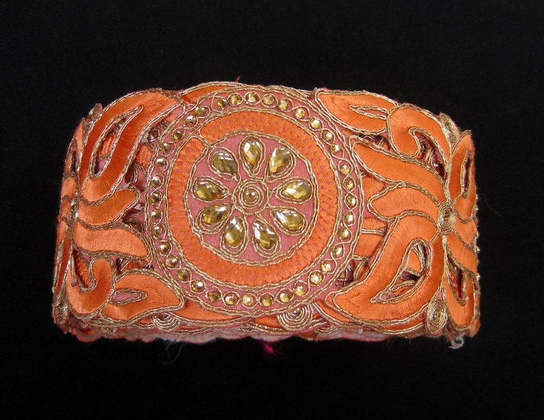 Op de afbeelding: Oranje stoffen bies met gouden borduurwerk en strass. De bies heeft een bloemmotief met een centraal cirkelmotief.