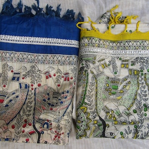 Puede incluir: Dos bufandas decorativas con intrincados diseños pintados a mano. Una es azul con ribete plateado y flecos, la otra es blanca con ribete amarillo y detalles verdes. Ambas presentan figuras detalladas y patrones florales.
