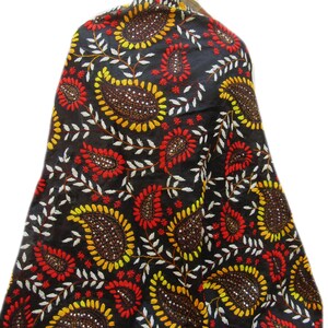 Op de afbeelding: Een zwart textiel met een herhalend paisley-patroon in geel, rood en bruin. De paisley-ontwerpen zijn omlijnd met wit, wat een opvallend contrast creëert. De stof is gedrapeerd, wat suggereert dat het een sjaal of omslagdoek kan zijn.