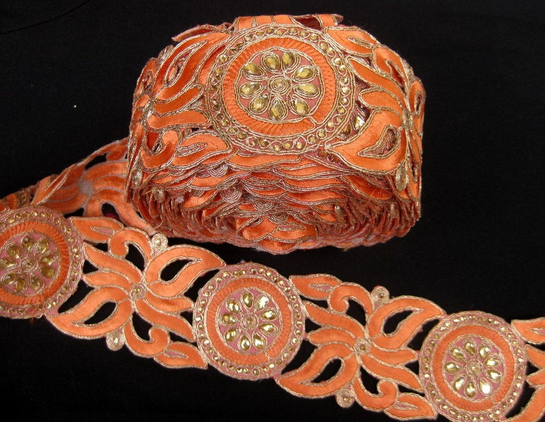 Op de afbeelding: Oranje geborduurde trim met een bloemenpatroon en gouden accenten. De trim heeft een herhalend patroon van cirkels met strass en uitgesneden bloemenvormen.