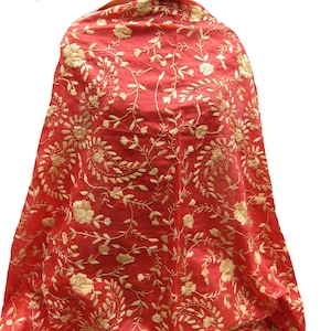 Könnte beinhalten: Ein leuchtend roter Stoffschal mit einem komplizierten, durchgehenden Stickmuster aus metallischem Goldfaden. Das Design besteht aus sich wiederholenden Blumenmotiven und geschwungenen Paisley-Formen, die die gesamte Oberfläche bedecken.