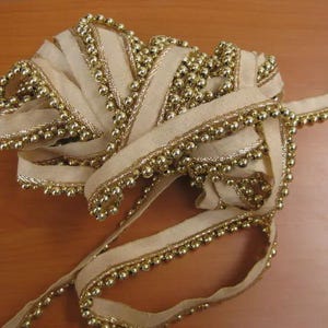 May include: A roll of ivory-colored ribbon with gold beaded trim. The ribbon is loosely coiled, showcasing the texture and detail of the gold beads along the edges. The ribbon's design suggests its use in crafts or decorative projects.