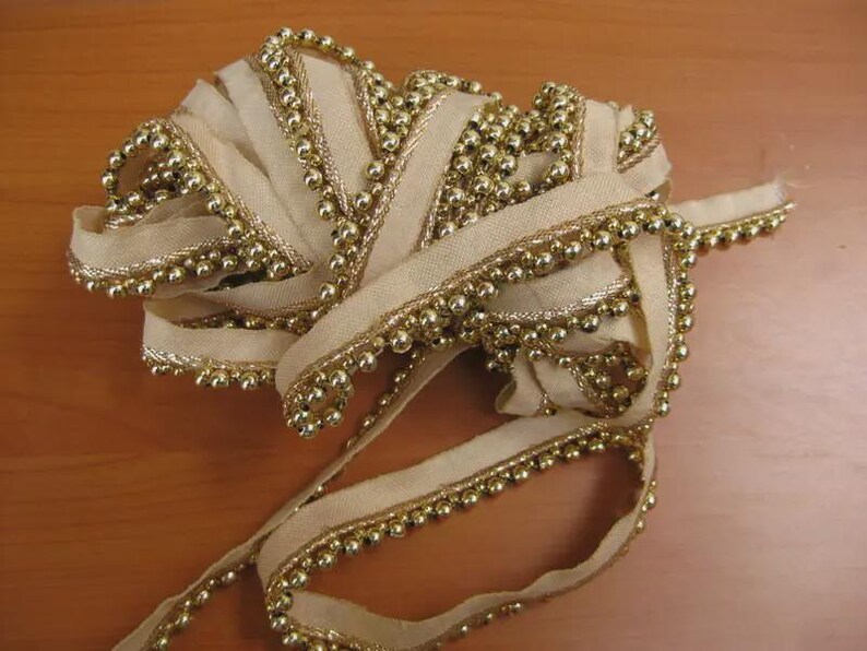 May include: A roll of ivory-colored ribbon with gold beaded trim. The ribbon is loosely coiled, showcasing the texture and detail of the gold beads along the edges. The ribbon's design suggests its use in crafts or decorative projects.