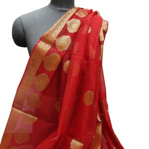 May include: A vibrant red scarf draped over a mannequin. The scarf features a gold border with a repeating geometric pattern and large gold circular designs. The fabric appears lightweight and flowing, suggesting it is made of silk or a similar material.