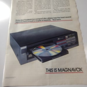 Puede incluir: Un reproductor de discos compactos negro con una cara plateada y un CD colorido en la bandeja. El texto "THIS IS MAGNAVOX" está impreso debajo del reproductor.