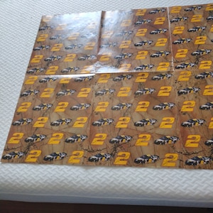Op de afbeelding: Een vel papier met een herhalend patroon van een zwarte raceauto met nummer 22 erop. De auto staat op een bruine en gele achtergrond met een marmerachtige textuur.