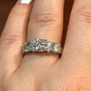 Pode incluir: Um anel de noivado de três pedras com uma pedra central grande e duas pedras menores de cada lado. O anel é engastado em ouro branco e apresenta um engaste pavé com diamantes menores.