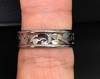 May include: A silver band ring with a decorative, intricate, cutout design. The ring is worn on a finger.