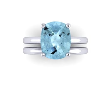 May include: A silver ring with a large, light blue aquamarine gemstone set in a cushion cut. The ring has a simple band and is designed to be worn as a wedding band.