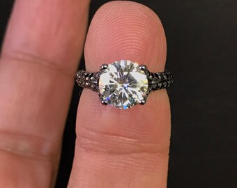 May include: A black ring with a large, clear, round gemstone. The ring is on a finger.