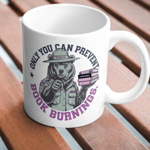 Pode incluir: Caneca de cerâmica branca com uma ilustração de desenho animado de um urso usando um chapéu de guarda florestal e segurando uma pilha de livros. O texto "Only you can prevent book burnings" está escrito em um círculo ao redor do urso.