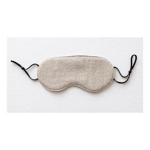 Op de afbeelding: Een beige slaapmasker van textiel met een textuur. Het masker heeft een voorgevormde vorm en is voorzien van zwarte, verstelbare bandjes. Ontworpen om licht te blokkeren.