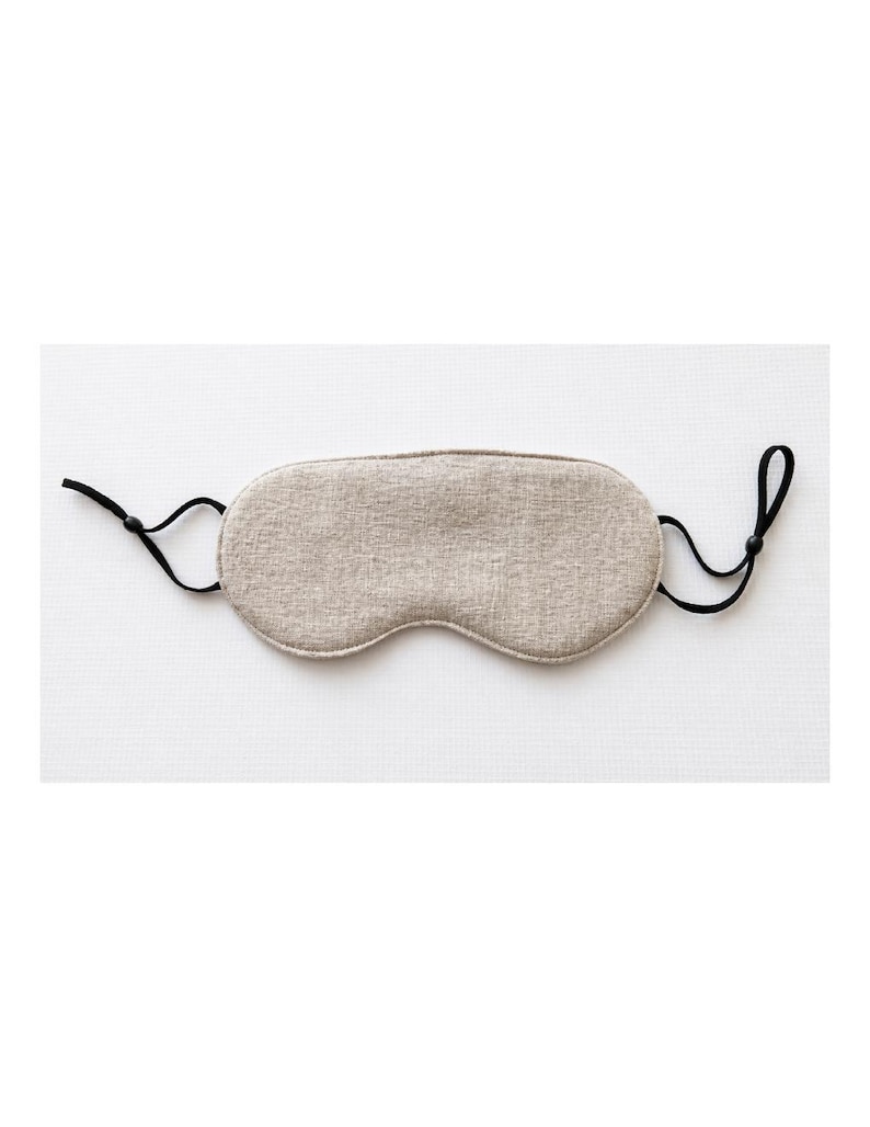 Op de afbeelding: Een beige slaapmasker van textiel met een textuur. Het masker heeft een voorgevormde vorm en is voorzien van zwarte, verstelbare bandjes. Ontworpen om licht te blokkeren.