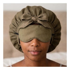 Op de afbeelding: Een olijfgroene slaapset met een slaapmasker en een muts met een strik. Het slaapmasker bedekt de ogen en de muts bedekt het hoofd. De set is gemaakt van een zachte, getextureerde stof.