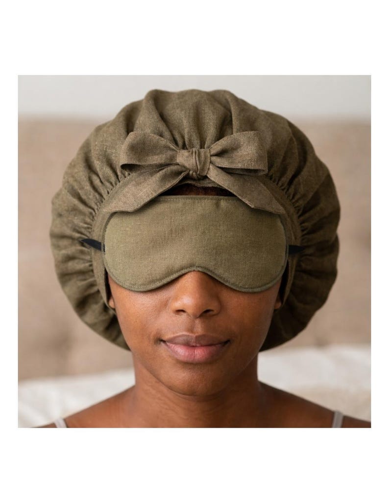 Op de afbeelding: Een olijfgroene slaapset met een slaapmasker en een muts met een strik. Het slaapmasker bedekt de ogen en de muts bedekt het hoofd. De set is gemaakt van een zachte, getextureerde stof.