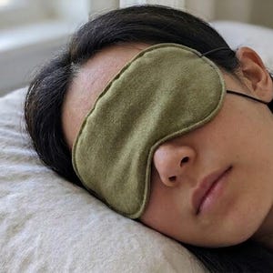 Op de afbeelding: Een groene slaapmasker van zacht, getextureerd materiaal. De persoon draagt het masker en ligt op een kussen. Het masker wordt vastgezet met een dunne zwarte elastische band. De persoon lijkt te slapen.