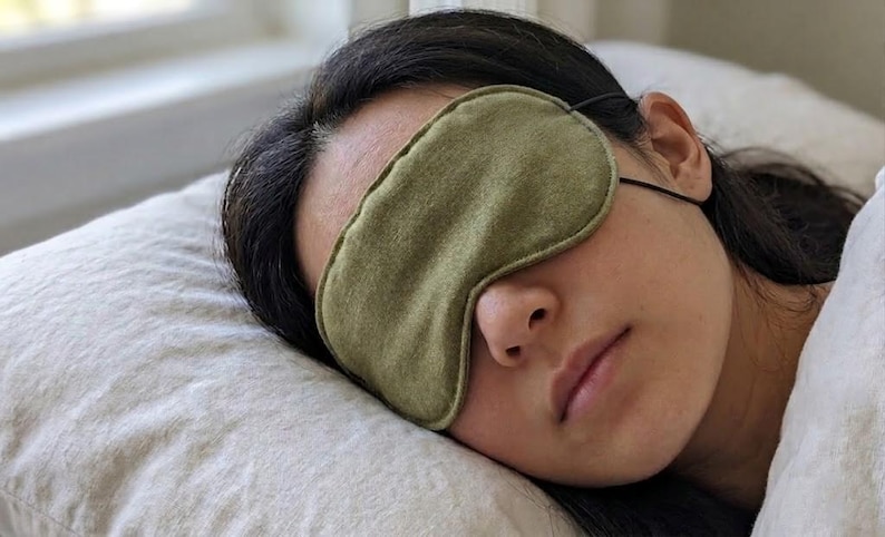 Op de afbeelding: Een groene slaapmasker van zacht, getextureerd materiaal. De persoon draagt het masker en ligt op een kussen. Het masker wordt vastgezet met een dunne zwarte elastische band. De persoon lijkt te slapen.