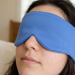 May include: A close-up of a person wearing a blue sleep mask. The mask is made of a soft fabric and covers the eyes and forehead. The person is resting, with their eyes closed.