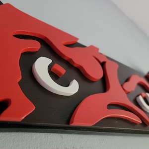 May include: A close-up of a wooden wall art piece with a stylized design. The artwork features a bold red design against a black background, with white accents. The piece is mounted on a wall.