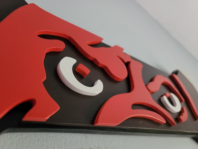 May include: A close-up of a wooden wall art piece with a stylized design. The artwork features a bold red design against a black background, with white accents. The piece is mounted on a wall.