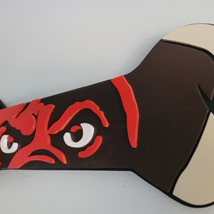 May include: A dog bone-shaped wall decoration featuring a stylized dog face. The dog's face is rendered in red and white against a dark brown background. The bone ends are beige, and the edges are black.
