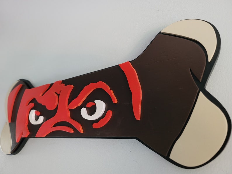 May include: A dog bone-shaped wall decoration featuring a stylized dog face. The dog's face is rendered in red and white against a dark brown background. The bone ends are beige, and the edges are black.