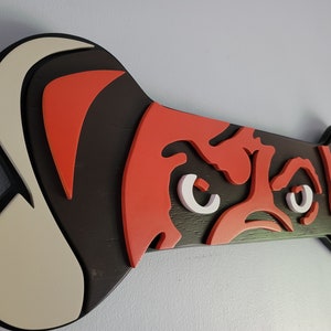 May include: A wooden dog bone-shaped wall art piece. The design features a stylized dog face in red, white, and black. The bone is approximately 12 inches long and 6 inches wide, with a black border.