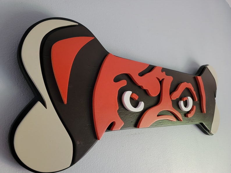 May include: A wooden dog bone-shaped wall art piece. The design features a stylized dog face in red, white, and black. The bone is approximately 12 inches long and 6 inches wide, with a black border.
