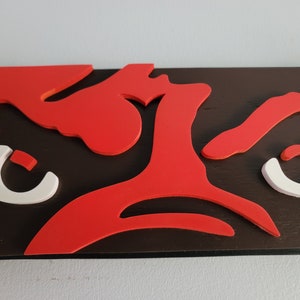 May include: A close-up of a wooden wall art piece featuring a bold design. The artwork showcases a dark brown base with vibrant red and white cut-out shapes. The shapes appear to be stylized eyes and other abstract forms.