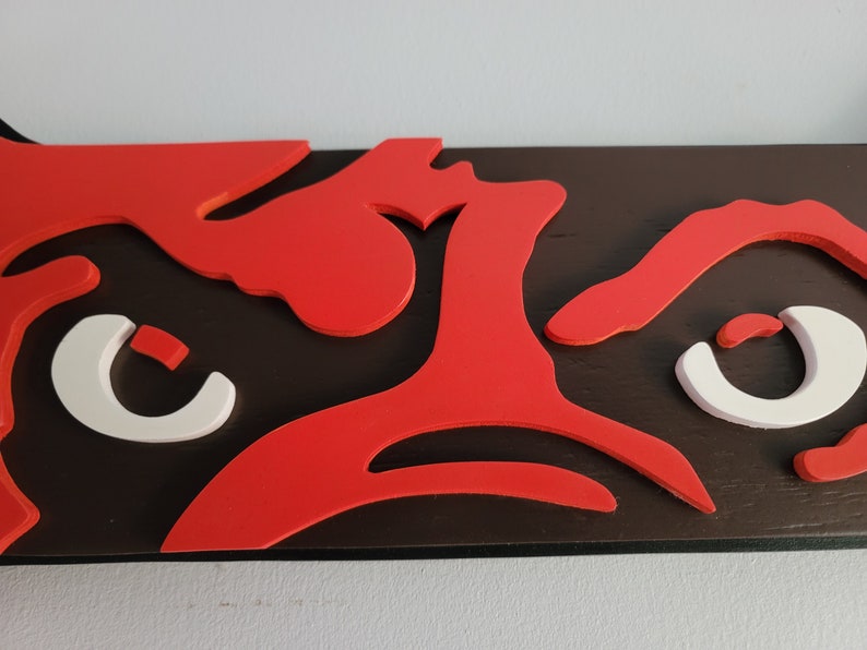 May include: A close-up of a wooden wall art piece featuring a bold design. The artwork showcases a dark brown base with vibrant red and white cut-out shapes. The shapes appear to be stylized eyes and other abstract forms.