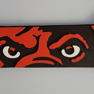 May include: A bone-shaped decorative item featuring a stylized dog face. The design incorporates orange, white, and black colors. The dog's eyes and facial features are prominently displayed on the dark brown bone.