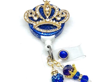 May include: A royal blue and gold badge reel with a crown design. The crown is gold with clear rhinestones. The reel has a blue glitter background and a dangling charm with blue beads and a small crown.