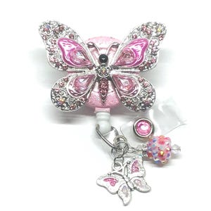 Puede incluir: Un portatarjetas en forma de mariposa rosa con detalles de pedrería. La mariposa tiene un contorno plateado y está adornada con pedrería rosa y transparente. Un pequeño dije de mariposa y detalles de cuentas cuelgan debajo.