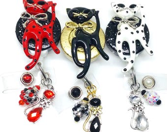 Può includere: Tre porta badge retrattili a tema gatto. Uno è rosso a pois neri, un altro è nero e il terzo è bianco a pois neri. Ogni gatto ha un fiocco e un ciondolo decorativo.