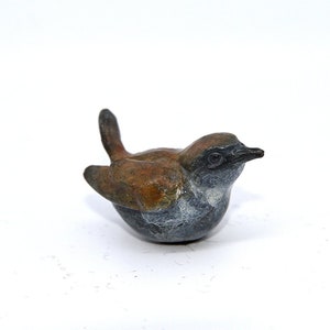 Könnte beinhalten: Eine kleine Bronzeskulptur eines Vogels mit braunen und grauen Federn. Der Vogel sitzt auf einer weißen Oberfläche.