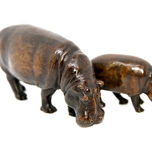 May include: Two bronze hippo figurines. The larger hippo is in front, with the smaller one behind it. Both hippos are facing down, as if grazing. The figurines have a dark brown colour and are set against a white background.
