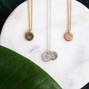 May include: Three gold necklaces with initial pendants on a white marble surface. The pendants are round and feature the letters "M", "D", and "B".
