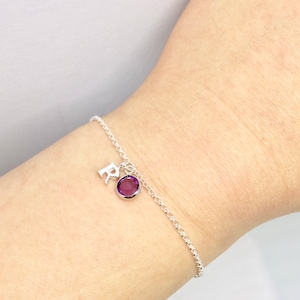 May include: A silver chain bracelet with a small purple gemstone and a silver charm with the letter "R" hanging from it.