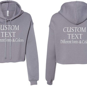 May include: A gray cropped hoodie with a drawstring hood. The hoodie has "CUSTOM TEXT" written on the front and back in white text. The text below says "Different fonts & Colors".