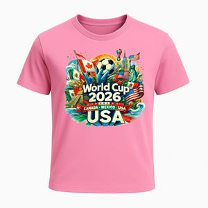 Puede incluir: Camiseta rosa con un dise&ntilde;o gr&aacute;fico colorido para la Copa del Mundo de 2026. El dise&ntilde;o incluye un bal&oacute;n de f&uacute;tbol, las banderas de Canad&aacute;, M&eacute;xico y EE. UU., y la Estatua de la Libertad.